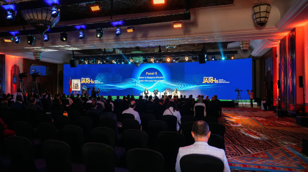 Vue d'ensemble d'une grande salle de conférence assombrie où de nombreux participants assistent à une table ronde. Sur la scène bien éclairée, quatre panélistes sont assis devant un écran géant bleu affichant le logo des JARHs et le thème « Panel 5 : Attirer la diaspora africaine : quels rôles pour les acteurs ». Des équipements de tournage vidéo sont visibles sur les côtés et un portrait officiel du Roi du Maroc est posé à gauche de l'estrade.