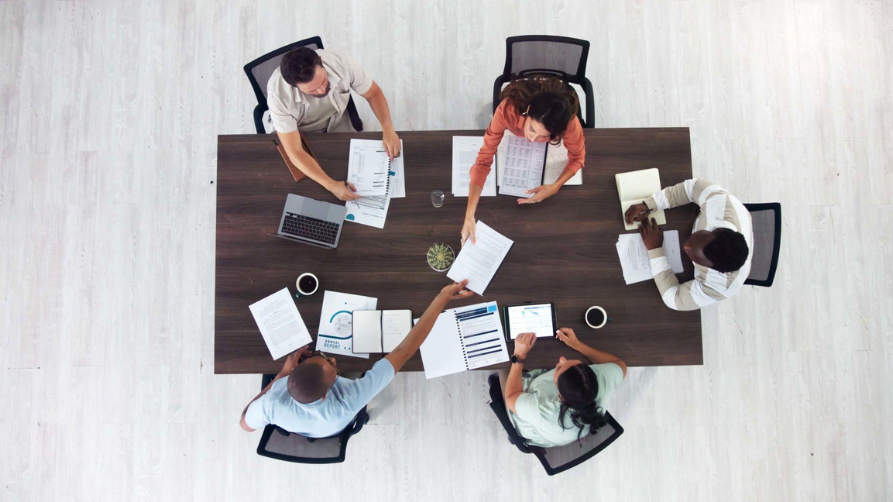 Une photo aérienne prise directement au-dessus d'une équipe de cinq professionnels divers (trois hommes et deux femmes) assis autour d'une grande table de conférence en bois sombre. Une femme en chemise rouille passe un document à un homme en chemise bleu clair. Le reste de l'équipe est engagé, regardant des documents de rapport, des ordinateurs portables et une tablette. Le sol en bois clair est visible autour de la table.