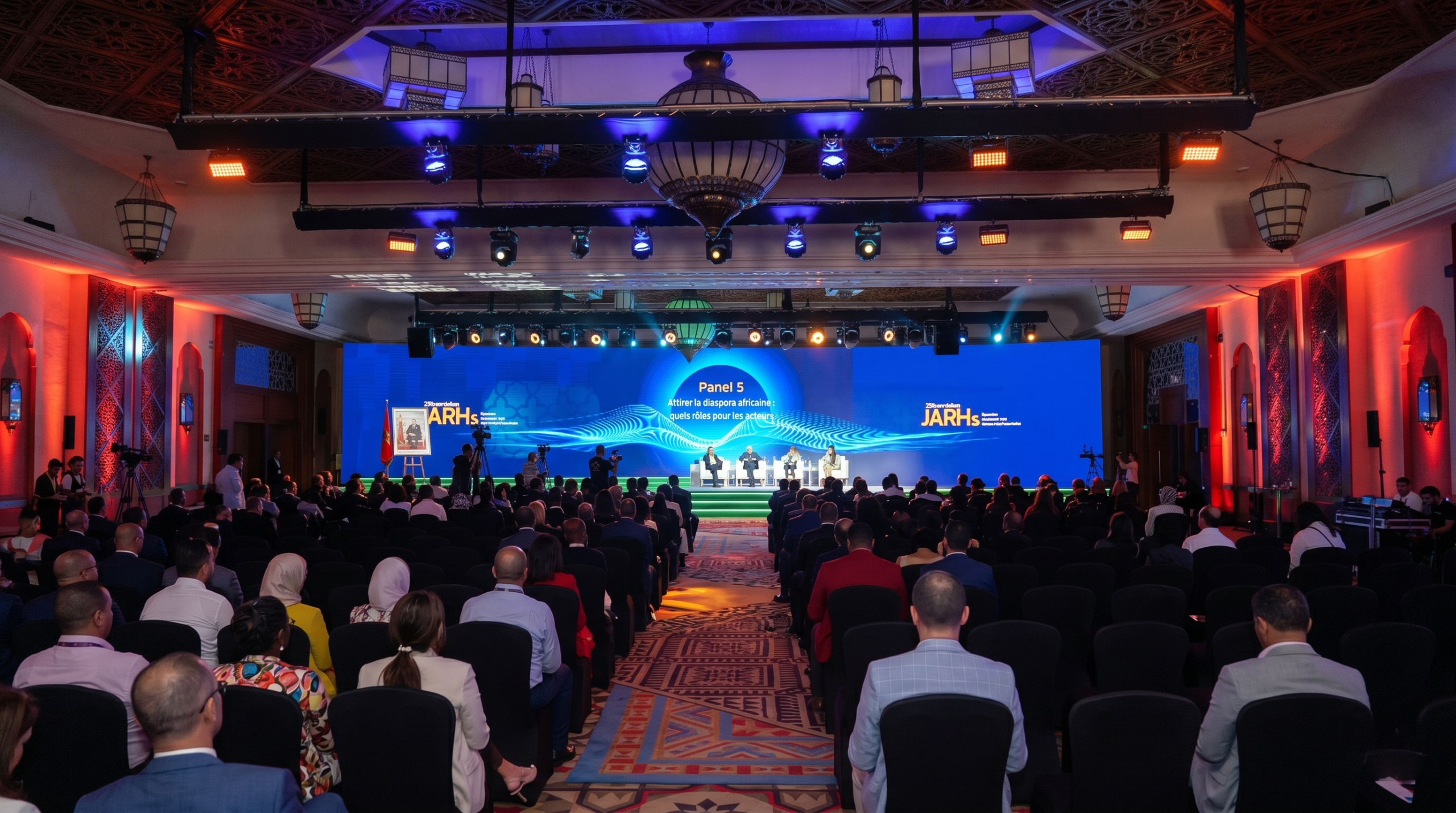 A wide-angle photograph of a large conference hall with an intricate Moroccan-style patterned ceiling and ornate lanterns, viewed from behind a seated audience. The main stage features a large LED screen displaying "Panel 5" and the French text "Attirer la diaspora africaine : quels rôles pour les acteurs," the "JARHs" logo, and a portrait. Four panelists are seated in front of the screen. The scene is dramatically lit with a mix of blue and orange light, and multiple camera operators are visible.