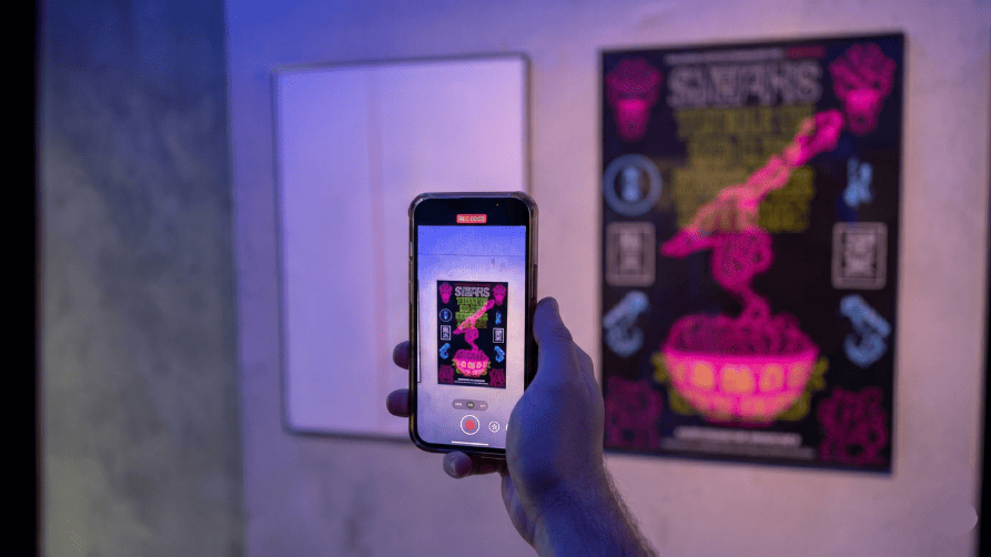 A close-up perspective of a person's hands holding a smartphone in a dimly lit room with purple ambient lighting. The phone's camera app is open and recording, focusing on a dark poster with vibrant neon pink and green designs. In the background, the actual neon poster is visible hanging on the wall to the right, next to a blurred, plain white framed canvas on the left.